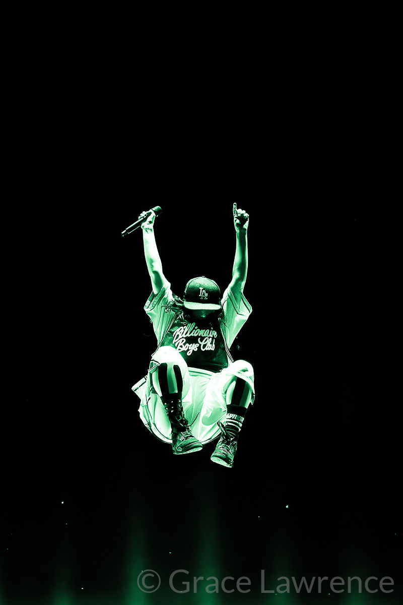 Billie Eilish mid-air with her arms up in the air and her legs tucked as she approaches the apex of her jump
